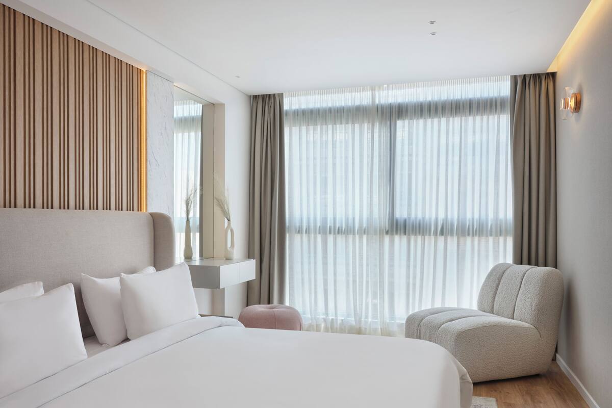 A modern hotel room with a large bed, a seating area, a vanity, and a large window with curtains.