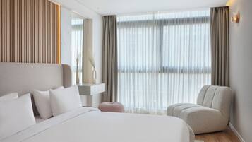 A modern hotel room with a large bed, a seating area, a vanity, and a large window with curtains.