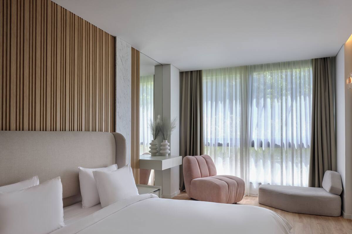 A modern hotel room with a large bed, a seating area, and a view of the outdoors through sheer curtains.
