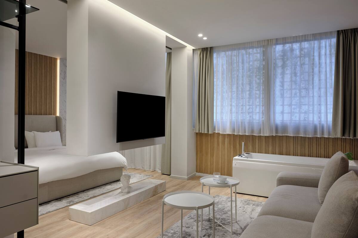 A modern hotel room with a bed, a TV mounted on the wall, a sofa, a small table, and a window with curtains.