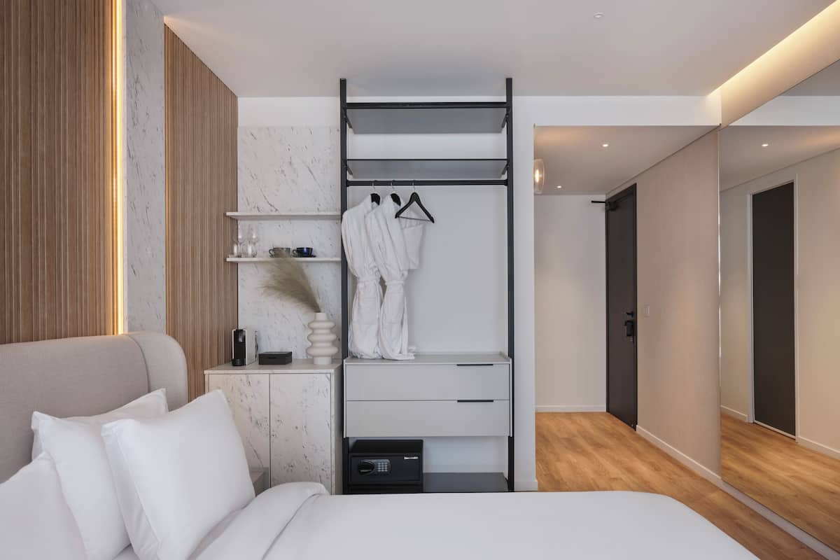 A modern hotel room with a bed, a bedside table with a lamp, a robe hanging on a rack, and a wardrobe with a mirror.