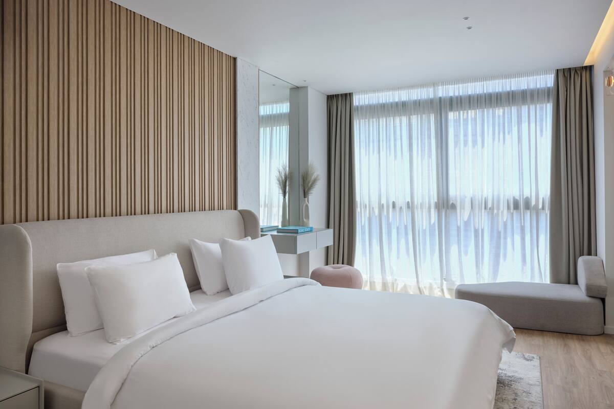 A modern hotel room with a large bed, a bedside table, a mirror, and a seating area with a chair.
