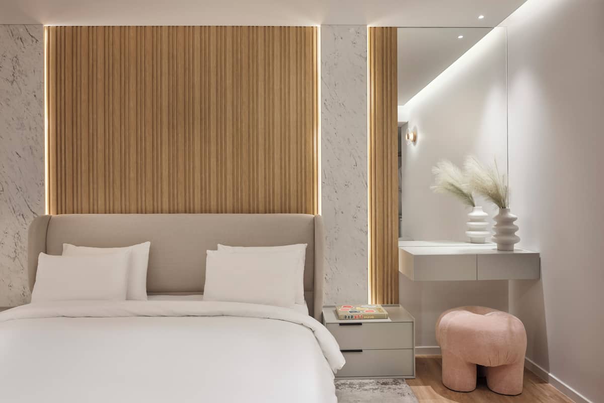 A modern bedroom with a large bed, a wooden panel wall, a bedside table with a book, and a pink ottoman.