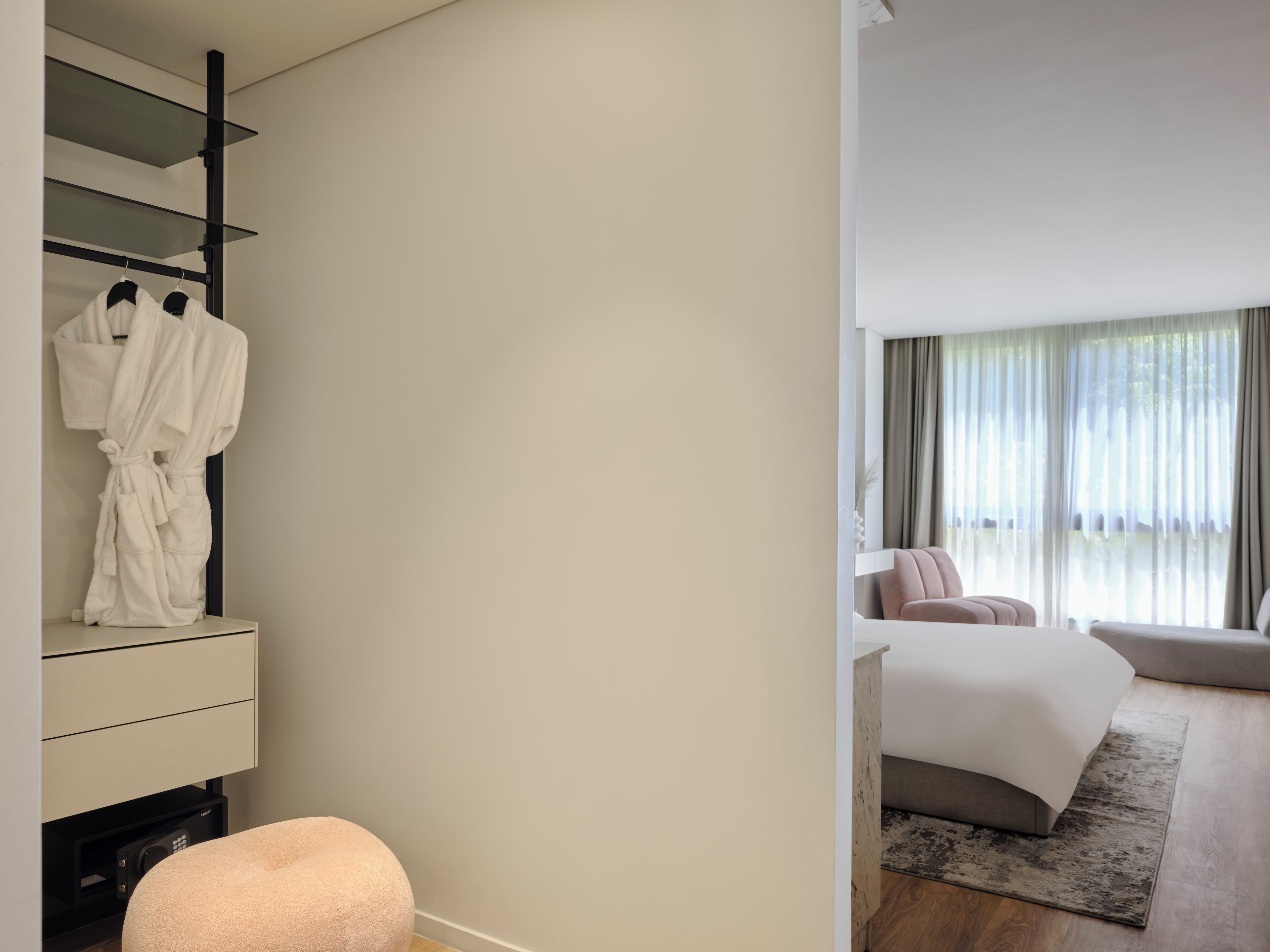 A modern hotel room with a built-in wardrobe, a bed with a grey headboard, and a large window with sheer curtains.