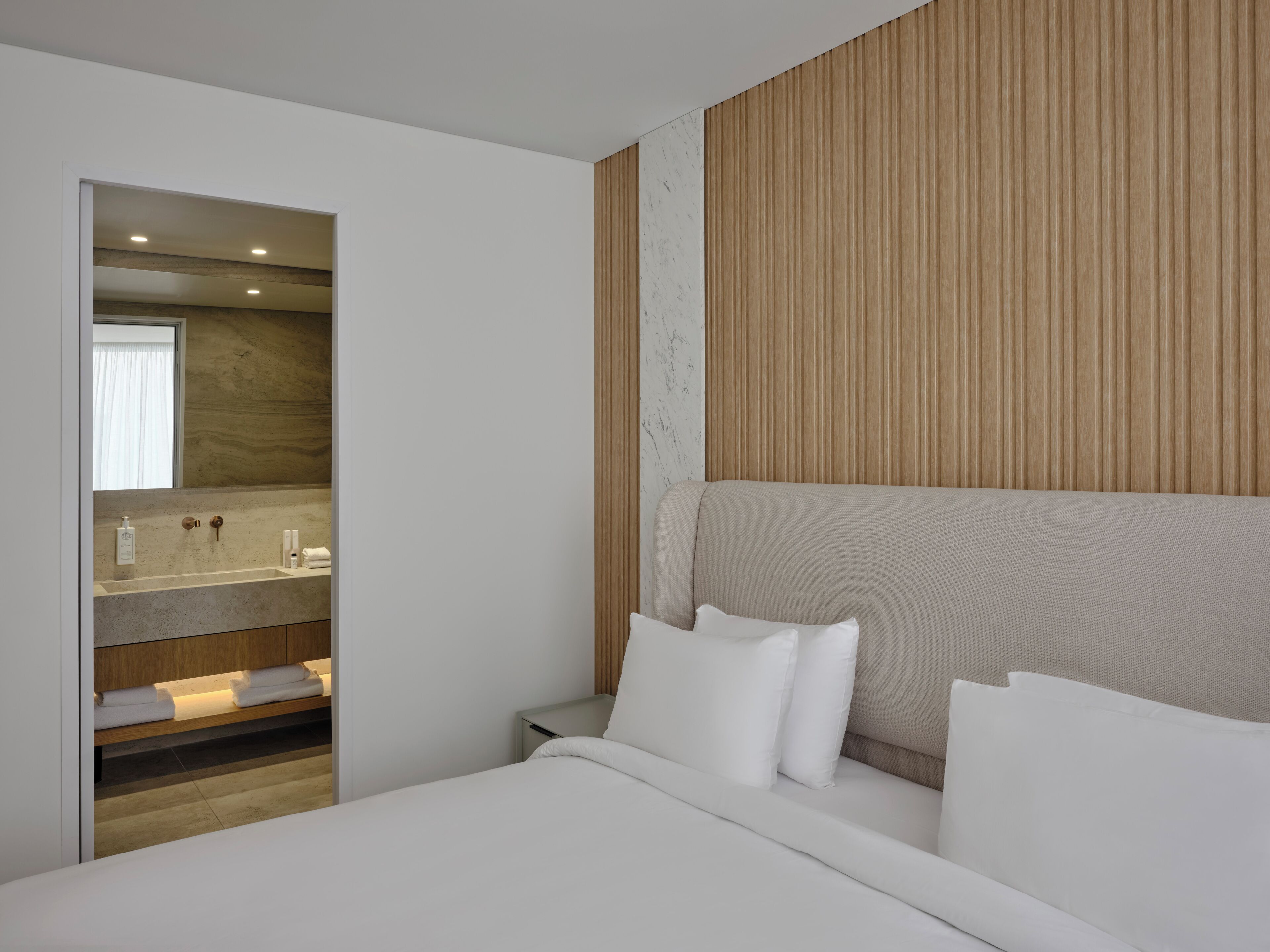 Quarto de hotel moderno com uma cama grande, painel de parede de madeira e banheiro visível através de uma porta aberta.
