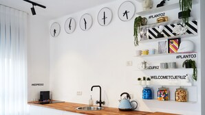 Eine moderne Küche mit Holzarbeitsplatte, Spüle, Wasserkocher und Gläsern mit Snacks. An den Wänden hängen Uhren und Regale mit Dekoration und Pflanzen.
