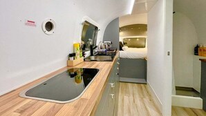 Una cocina compacta con pileta, cocina y alacenas, un pequeño comedor con mesa y sillas, y un dormitorio con cama y mesita de noche.