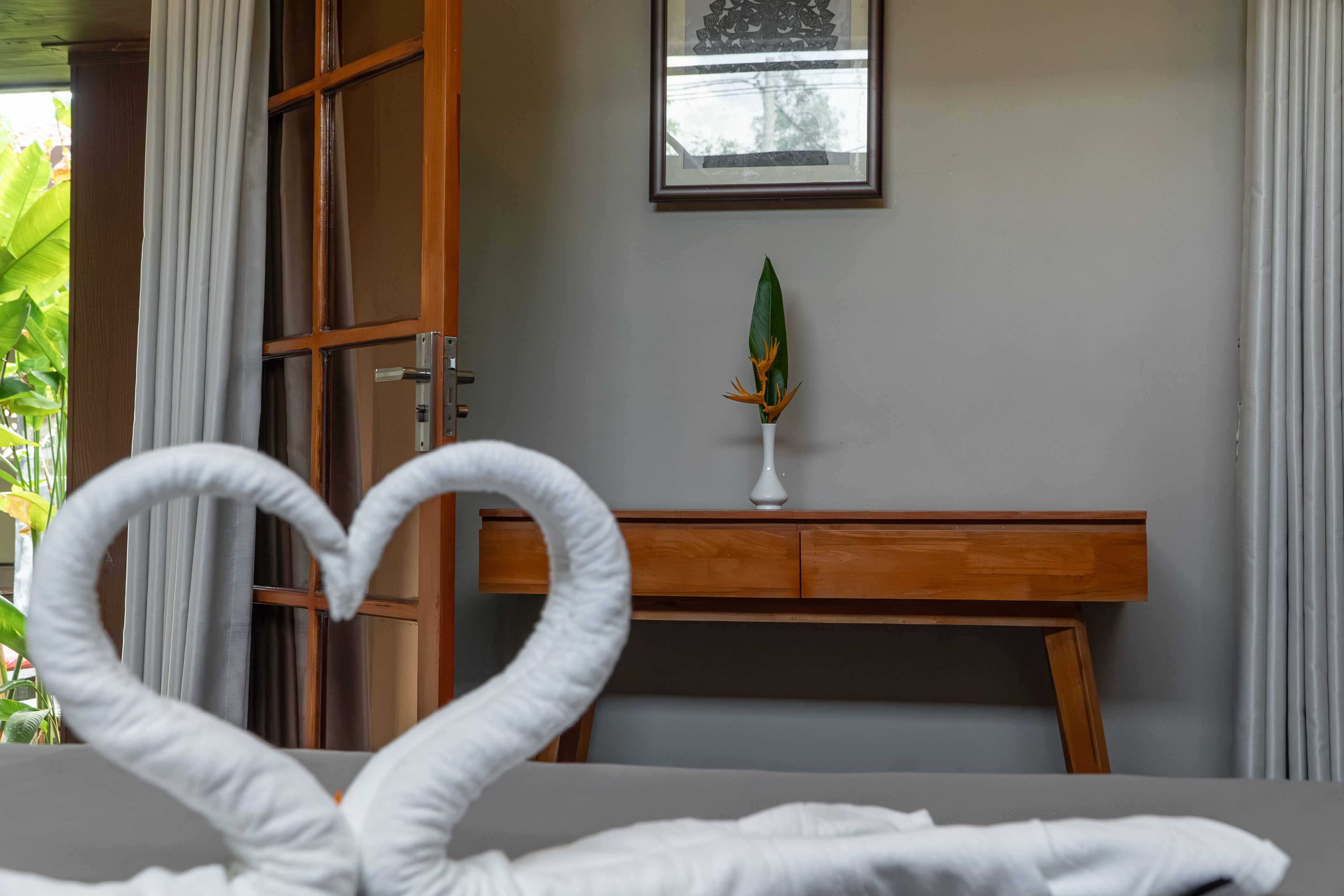 A hotel room with a bed, a wooden nightstand, a framed picture on the wall, and a potted plant.