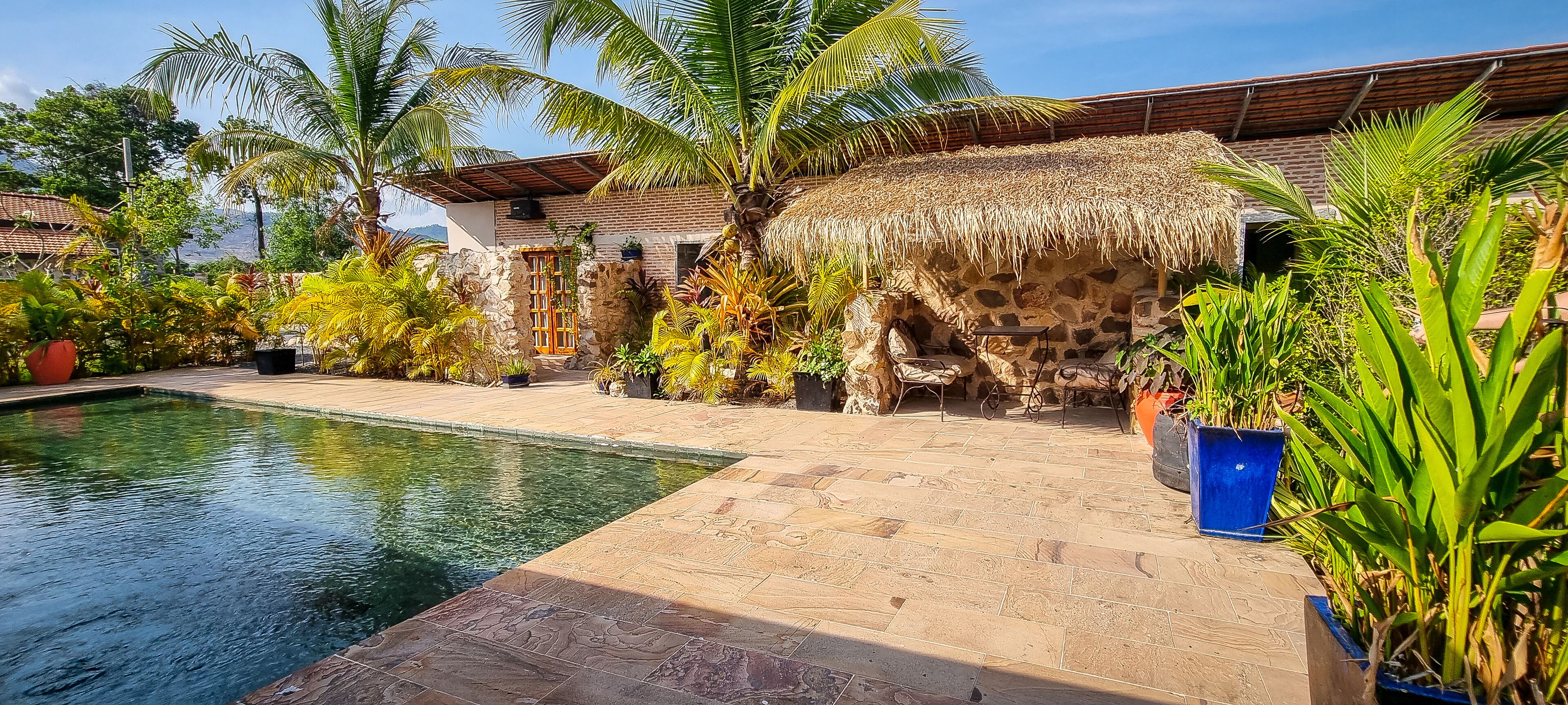 A pool area with a stone patio, surrounded by tropical plants and a thatched-roof structure.