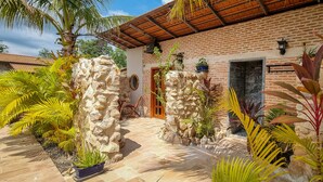 A brick building with a stone wall, a wooden door, and a tiled patio with tropical plants. - Villa Naya (Kampot)