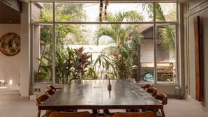 A dining area with a large table and chairs, a ceiling fan, and a view of greenery outside. - Villa Naya (Kampot)