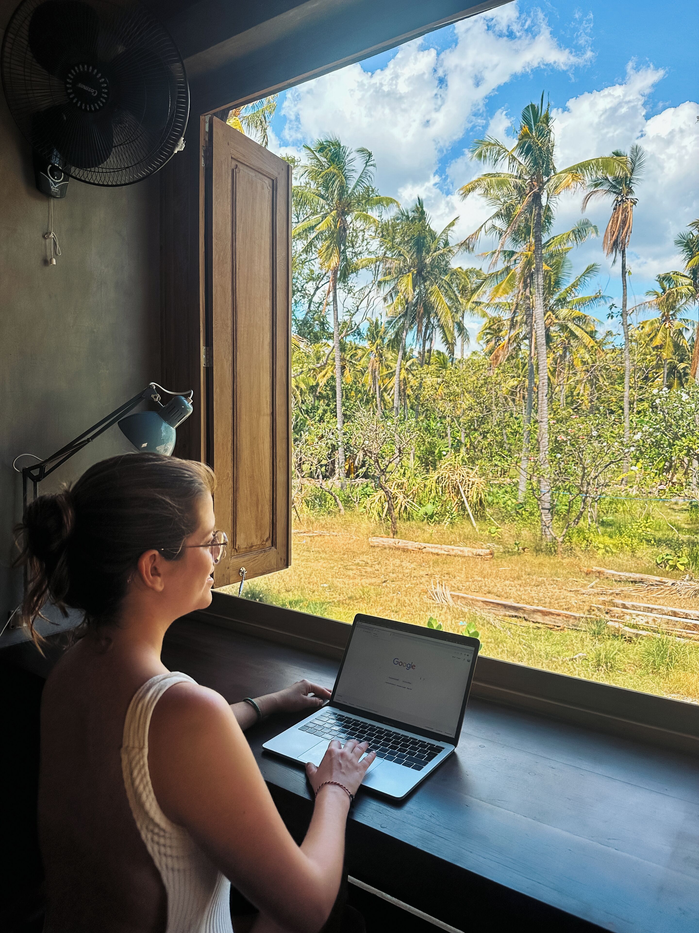 Une personne travaille sur un ordinateur portable, assise à un bureau en bois près d’une fenêtre qui offre une vue sur des palmiers et un ciel dégagé.