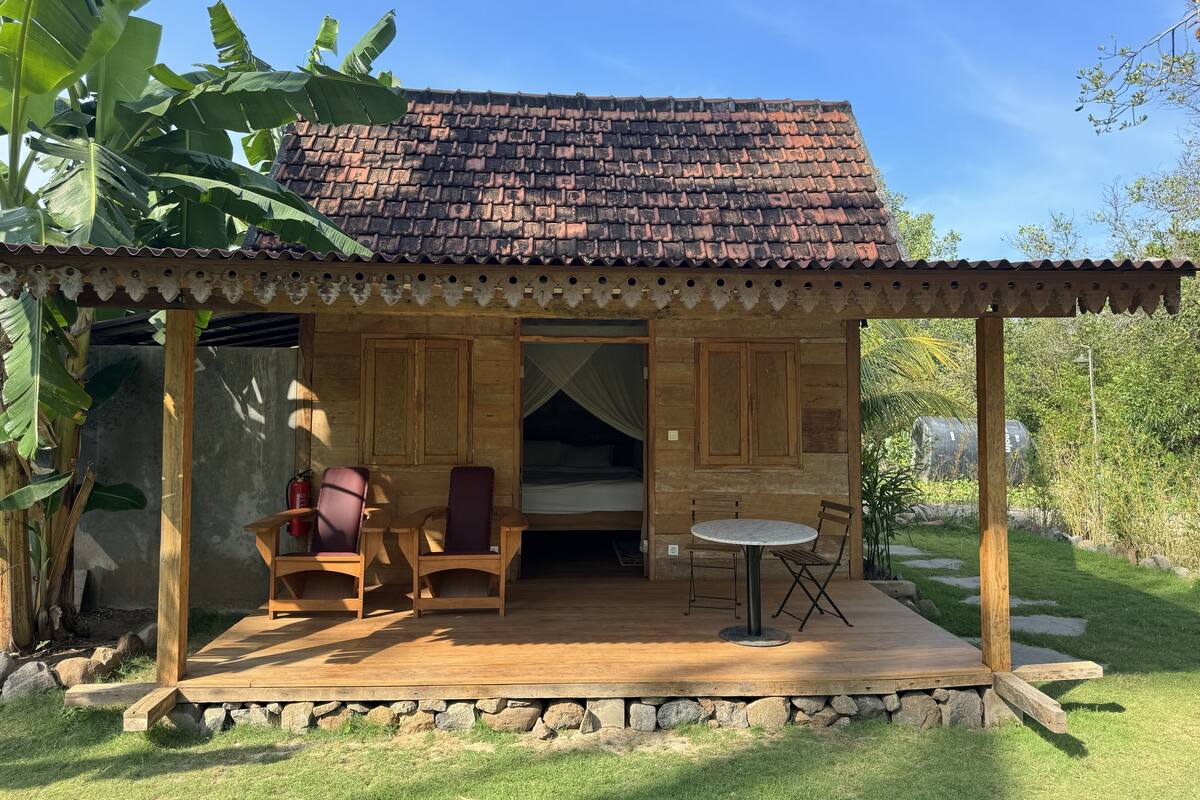 A wooden cabin with a tiled roof, a covered patio with chairs and a table, and a bed with a canopy.