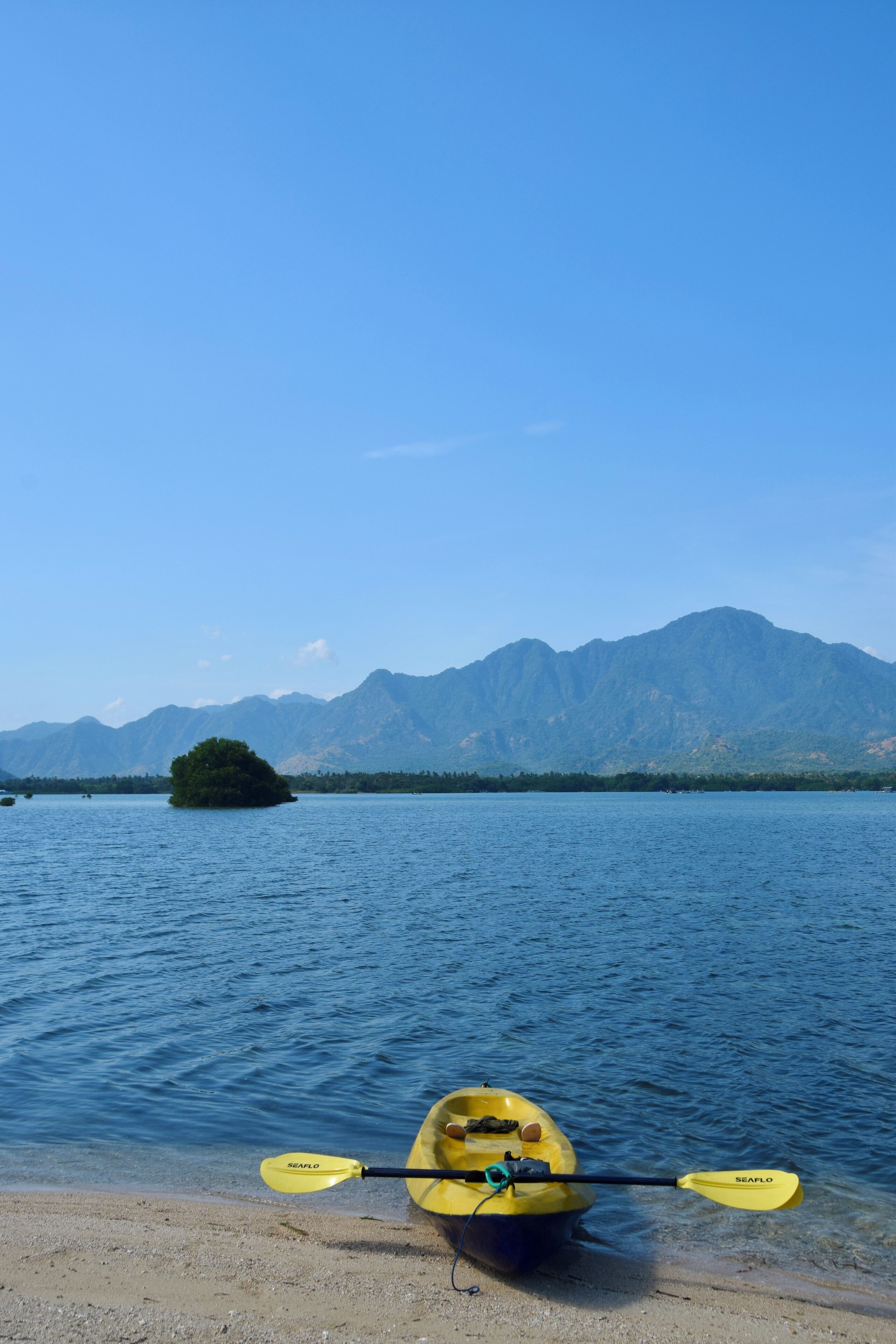 Una kayak con dos remos sobre una playa arenosa, un lago tranquilo y una cadena montañosa al fondo.