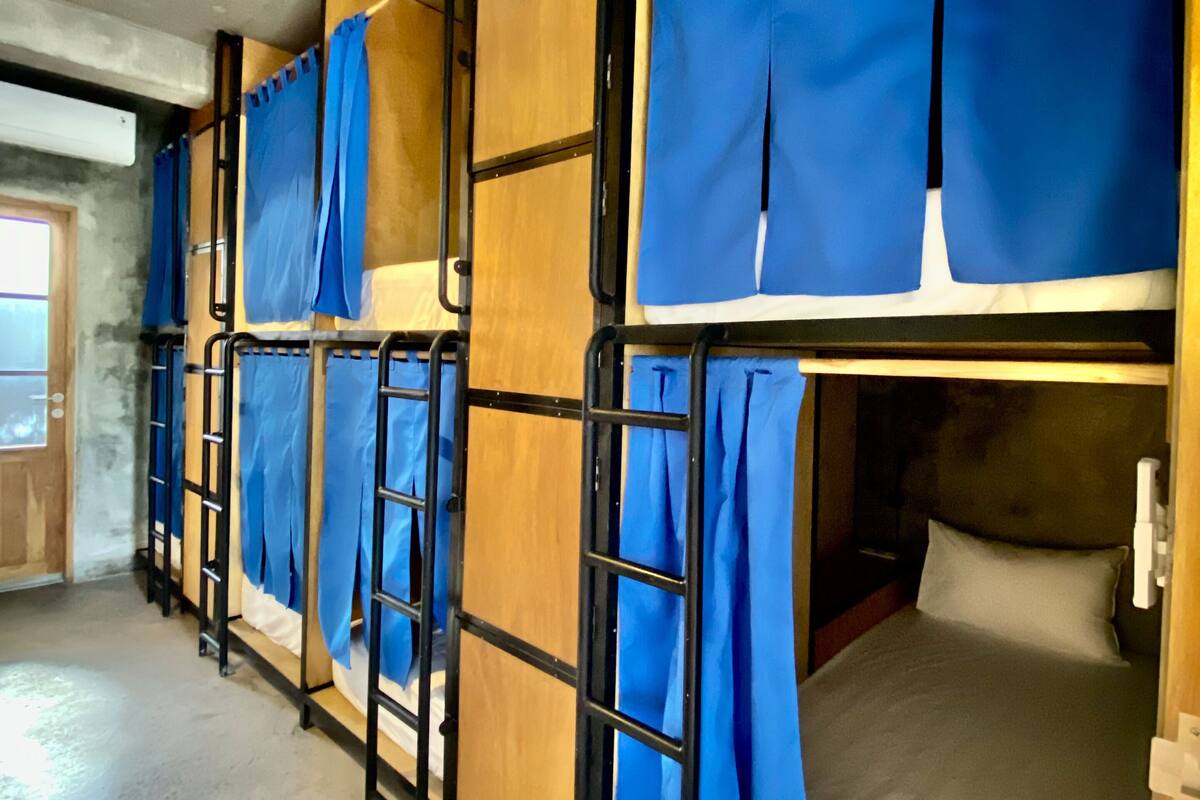 A dormitory room with bunk beds, each with blue curtains, wooden headboards, and a concrete floor.