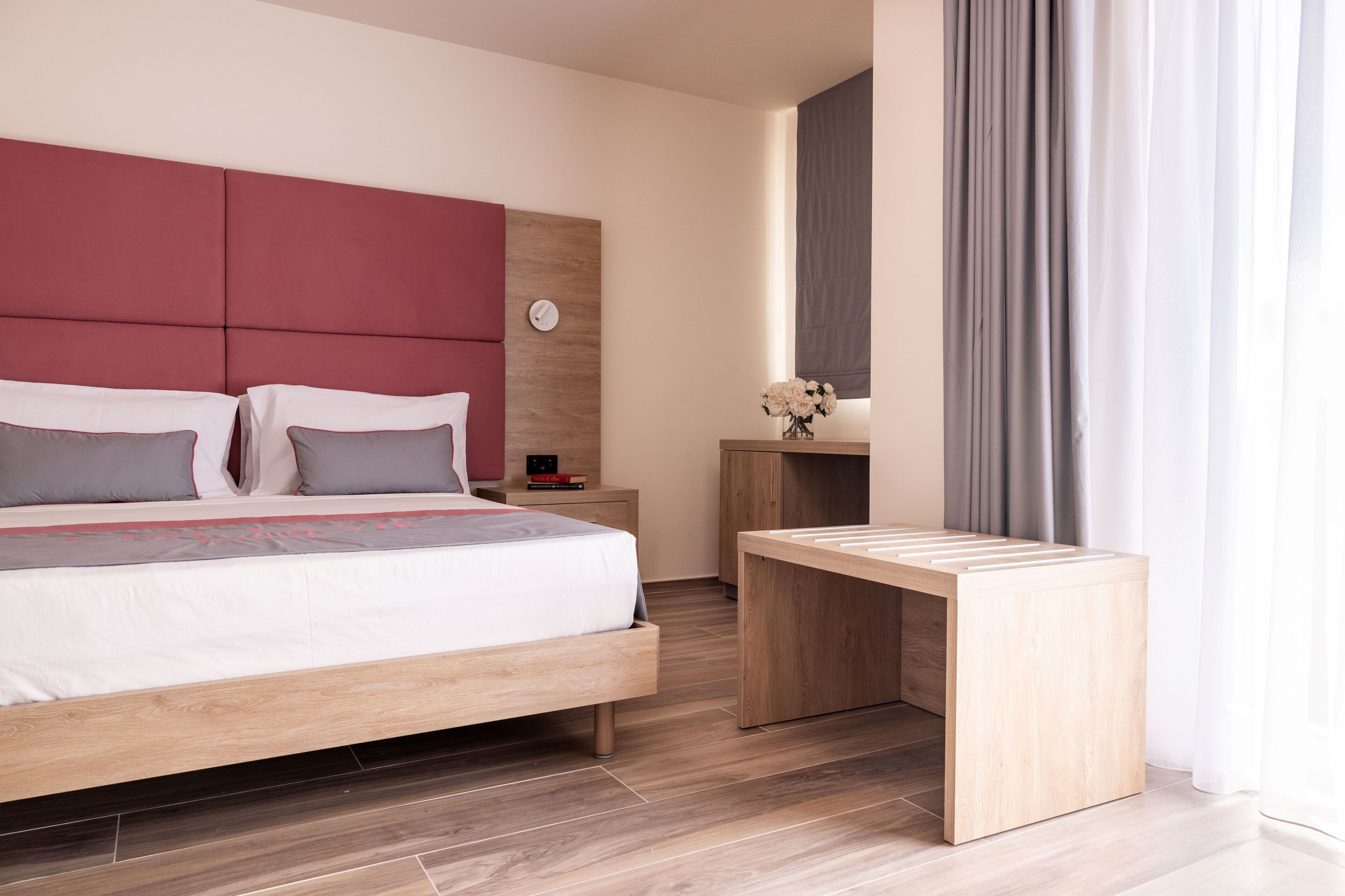 A modern hotel room with a large bed, a wooden nightstand, a small table with a vase of flowers, and a large window with curtains.