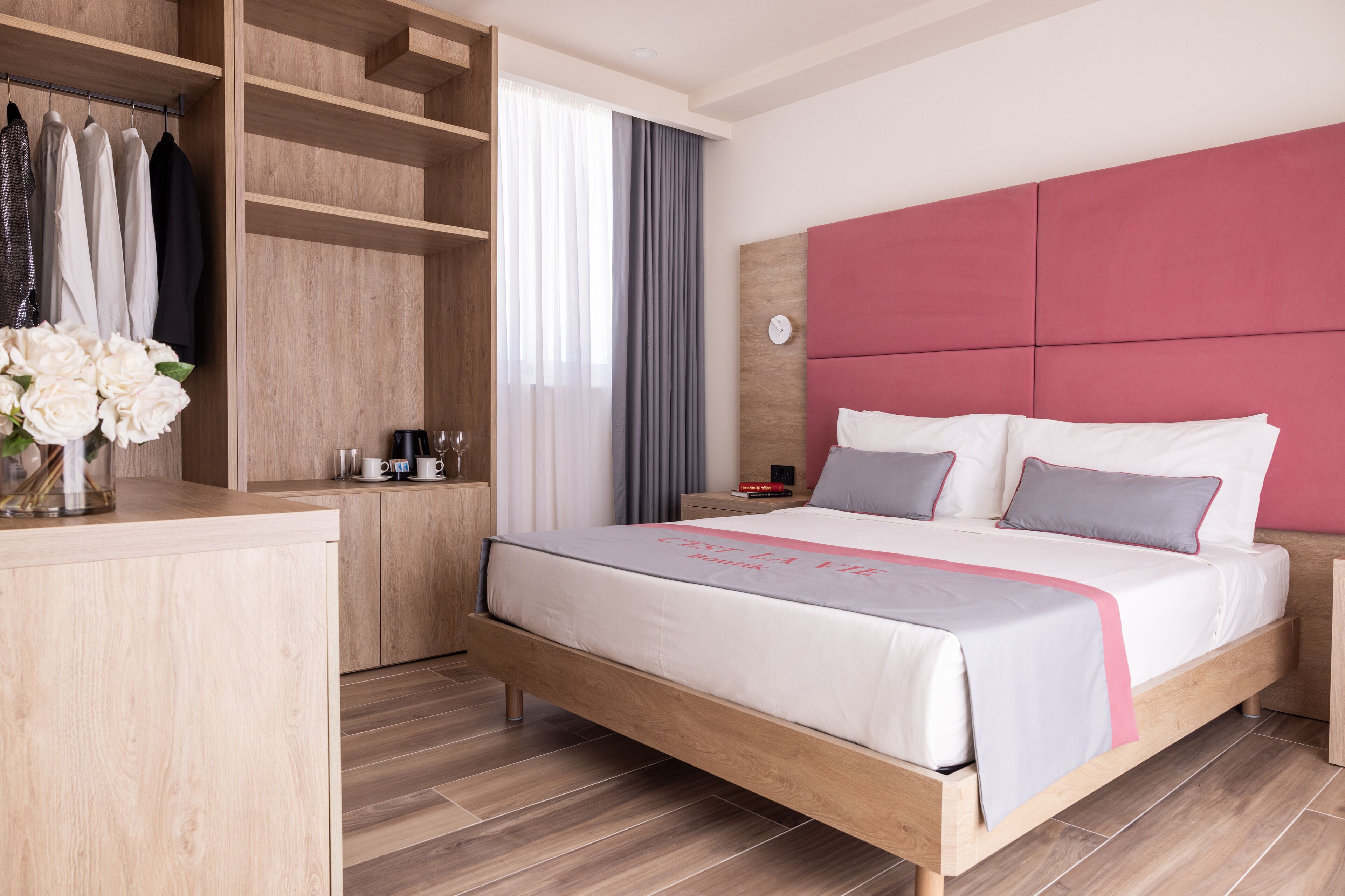 Moderni hotellihuone, jossa on puinen sängynrunko, vaaleanpunainen pääty ja sisäänrakennettu vaatekaappi, jossa on tilaa ripustettaville vaatteille.