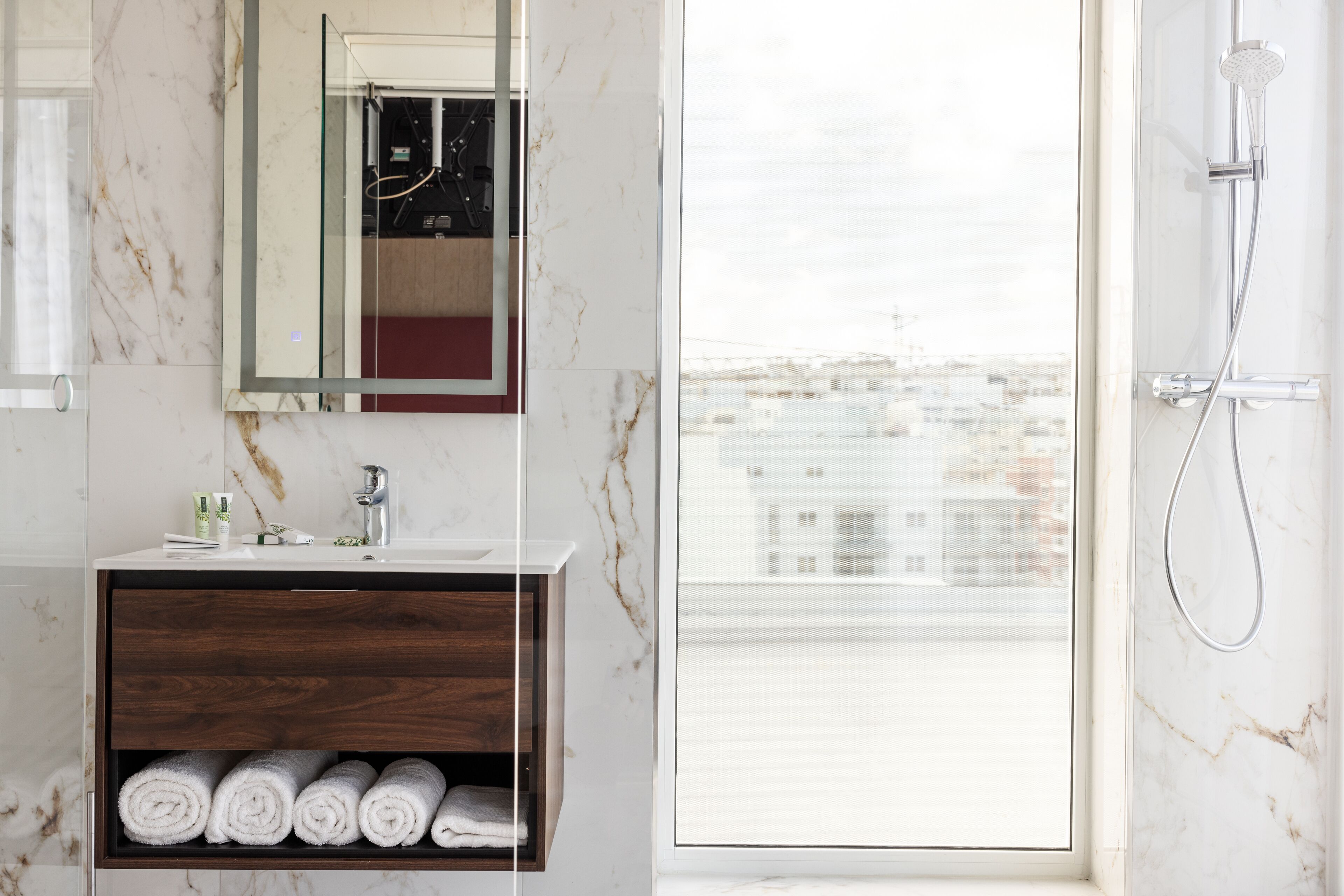 A bathroom with a marble wall, a wooden vanity, a mirror, a shower, and a view of buildings through a large window.
