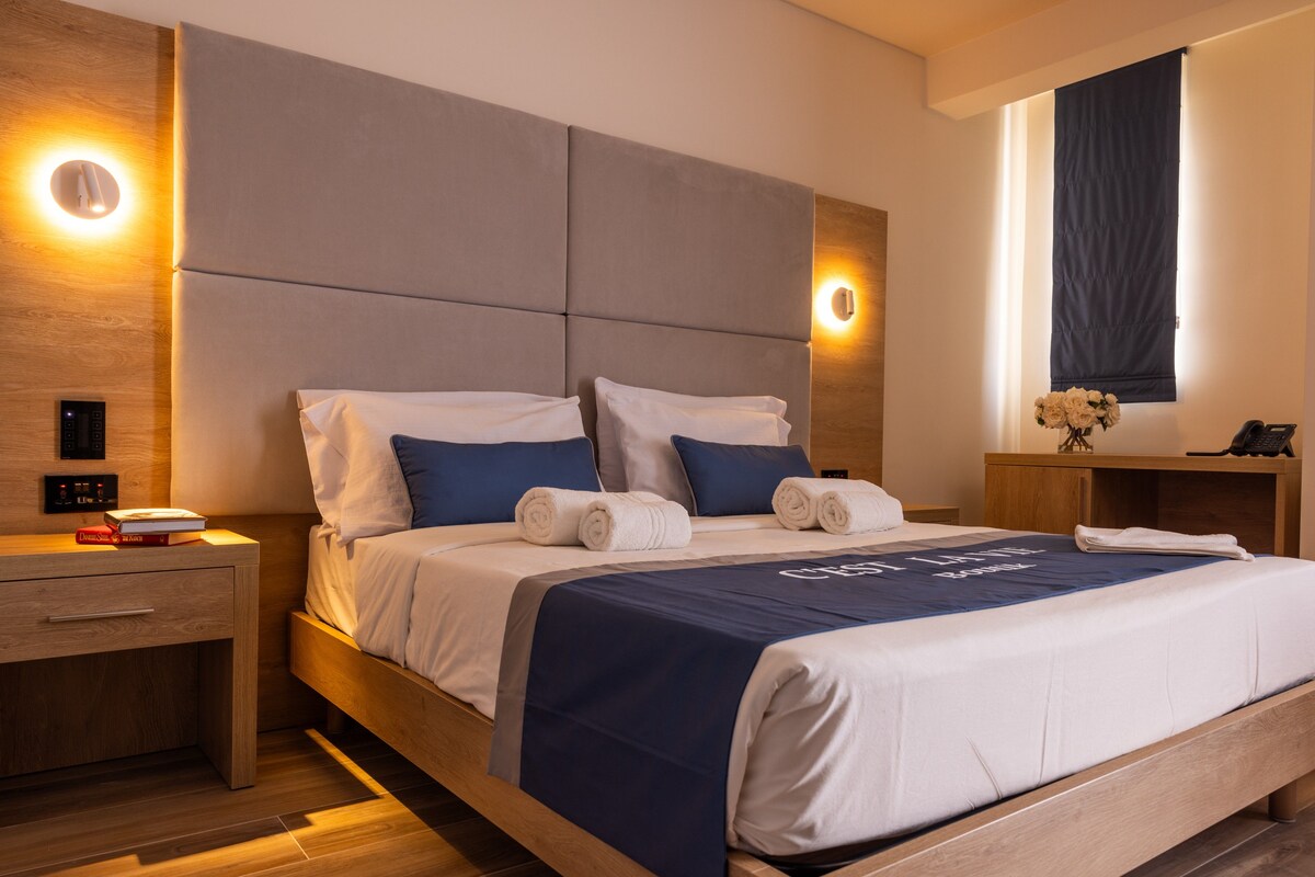 Una habitación de hotel moderna con una cama grande, cabecera de madera, mesitas de noche y una mesita de noche con un teléfono y libros.
