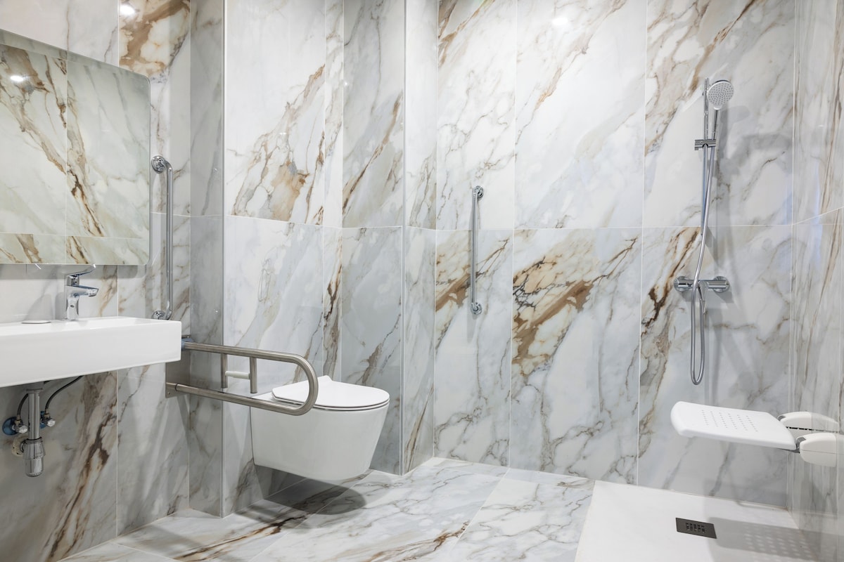 Un baño moderno con paredes de mármol, un lavabo blanco, un inodoro y una zona de ducha.