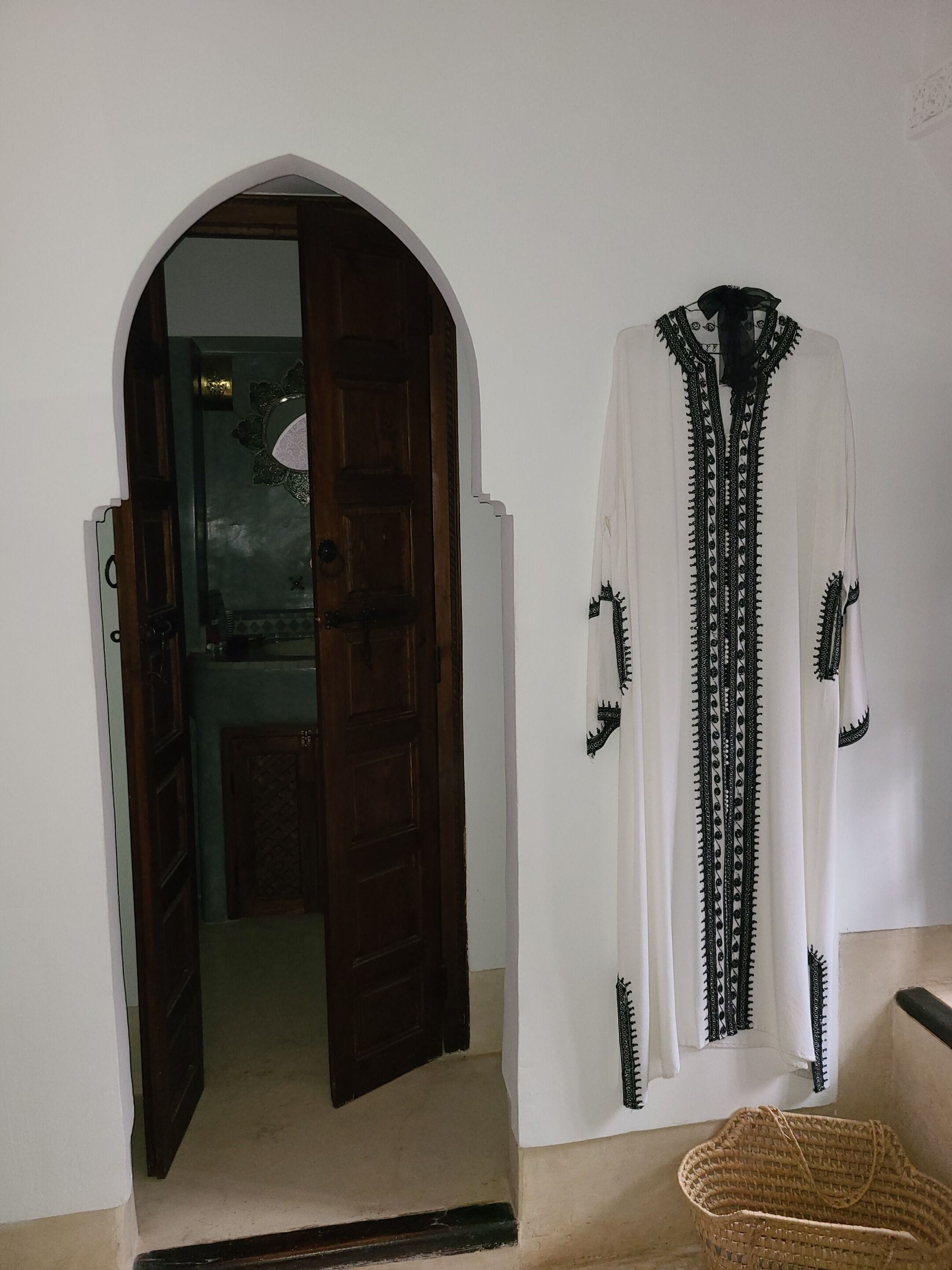 A doorway leading to a room with a traditional design, a white garment with black embroidery hanging on the wall, and a woven basket on the floor.