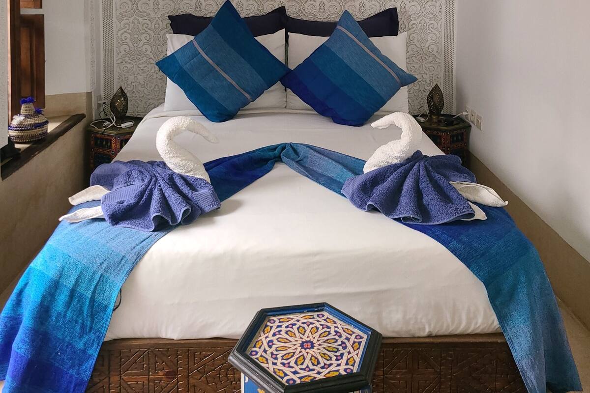 A bed with a white and blue patterned bedspread, two blue pillows, and a blue and white throw blanket.
