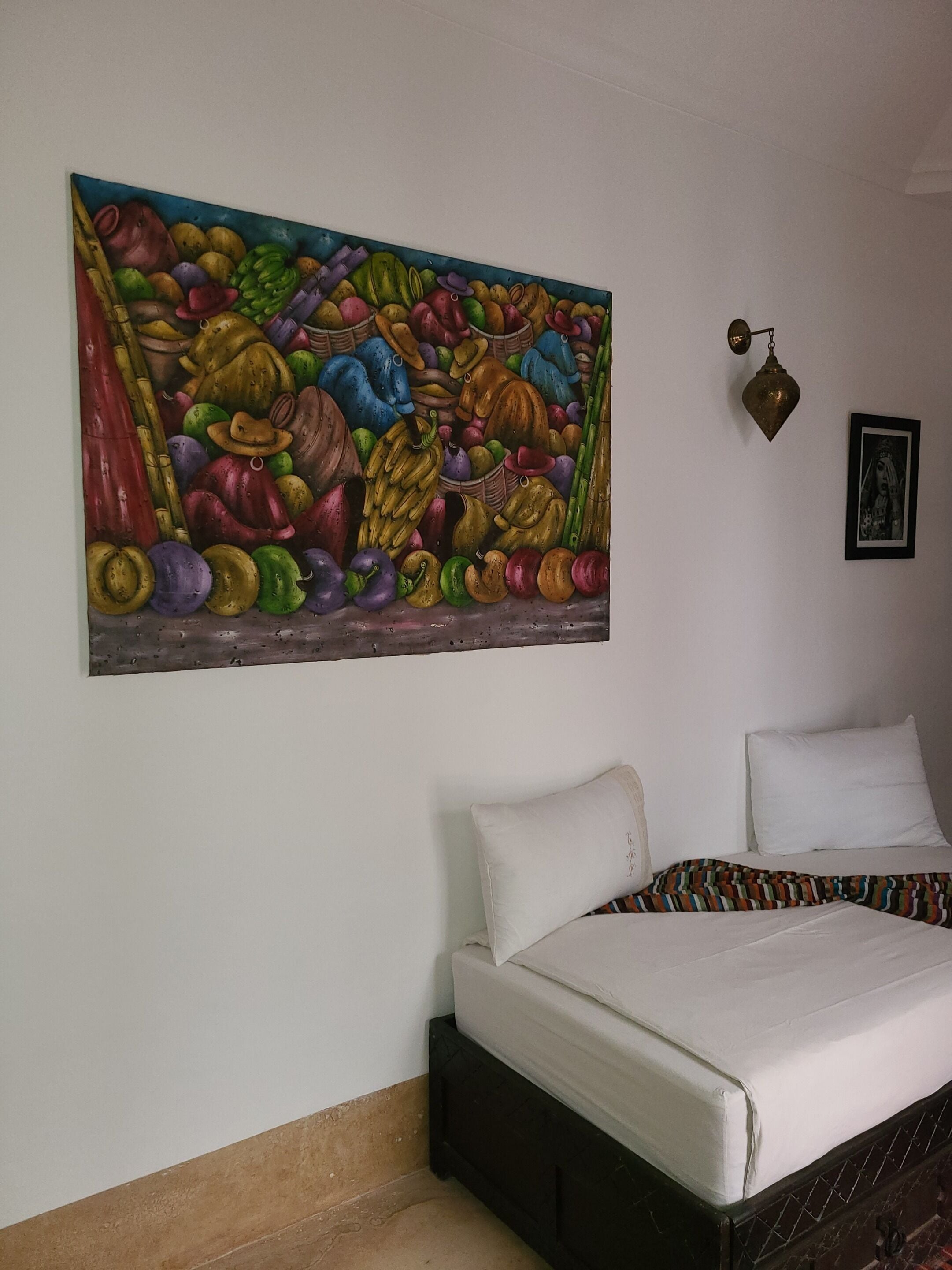 A room with a bed, a colorful painting on the wall, and a small hanging light fixture.