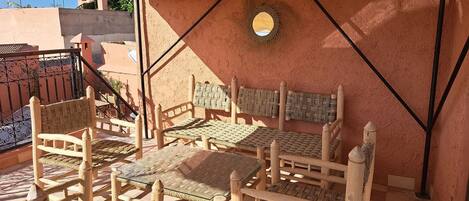 A sunlit outdoor seating area with wooden furniture and woven mats, a metal railing, and a textured wall.