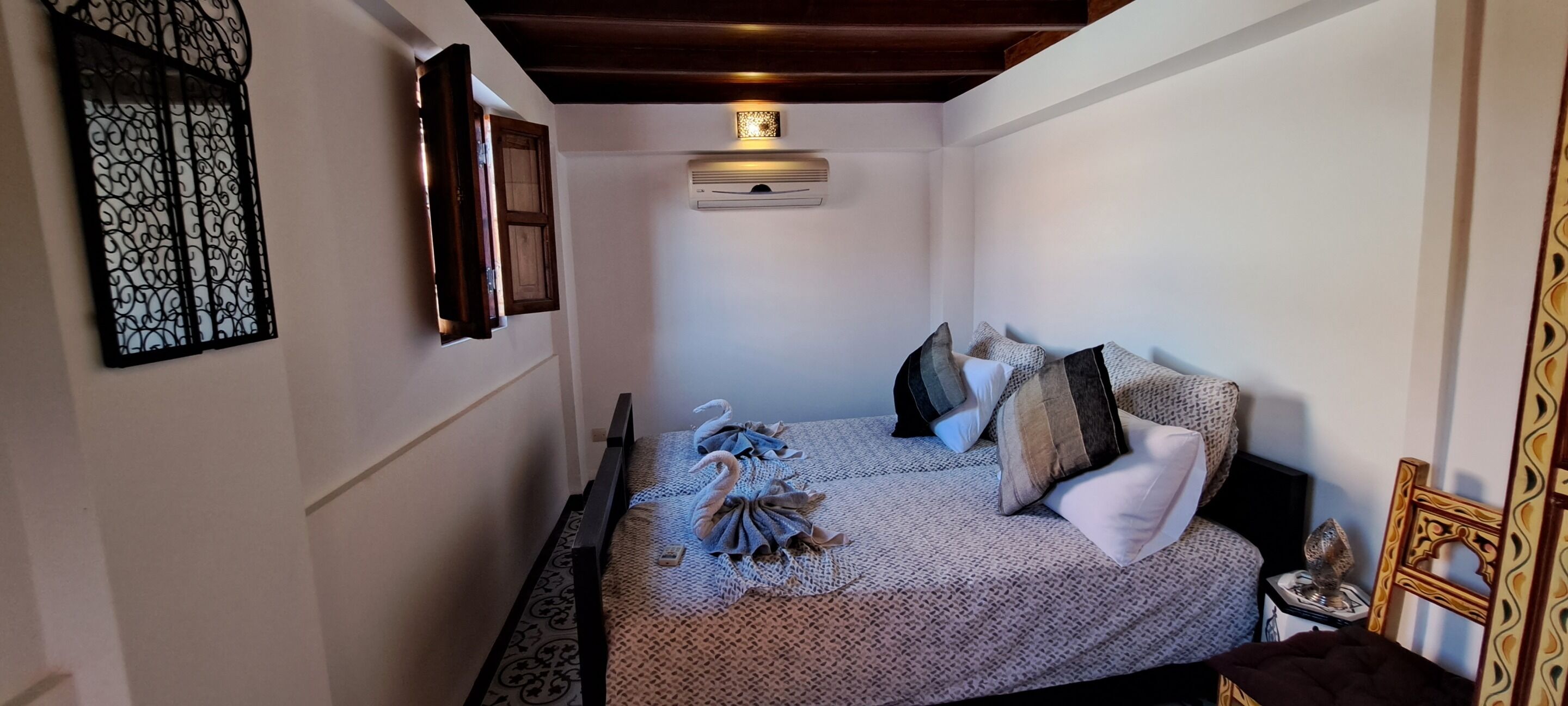 A narrow room with a bed, a wall-mounted air conditioner, a decorative wall piece, and a wooden ceiling.