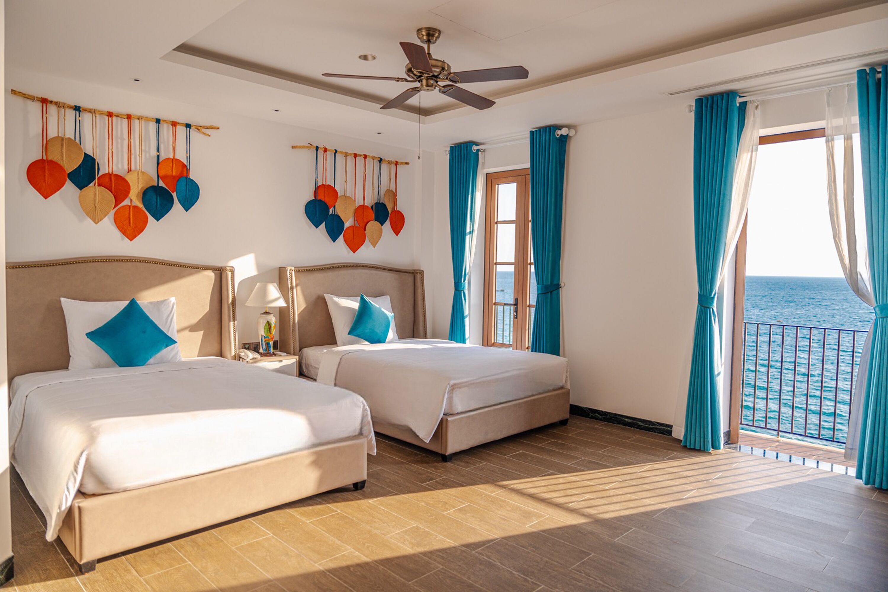 A hotel room with two beds, a ceiling fan, a view of the sea, and a balcony with curtains.