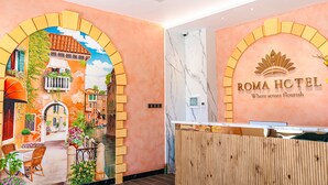 A hotel reception area with a mural of a street scene, a reception desk, and the hotel name 'ROMA HOTEL' on the wall.