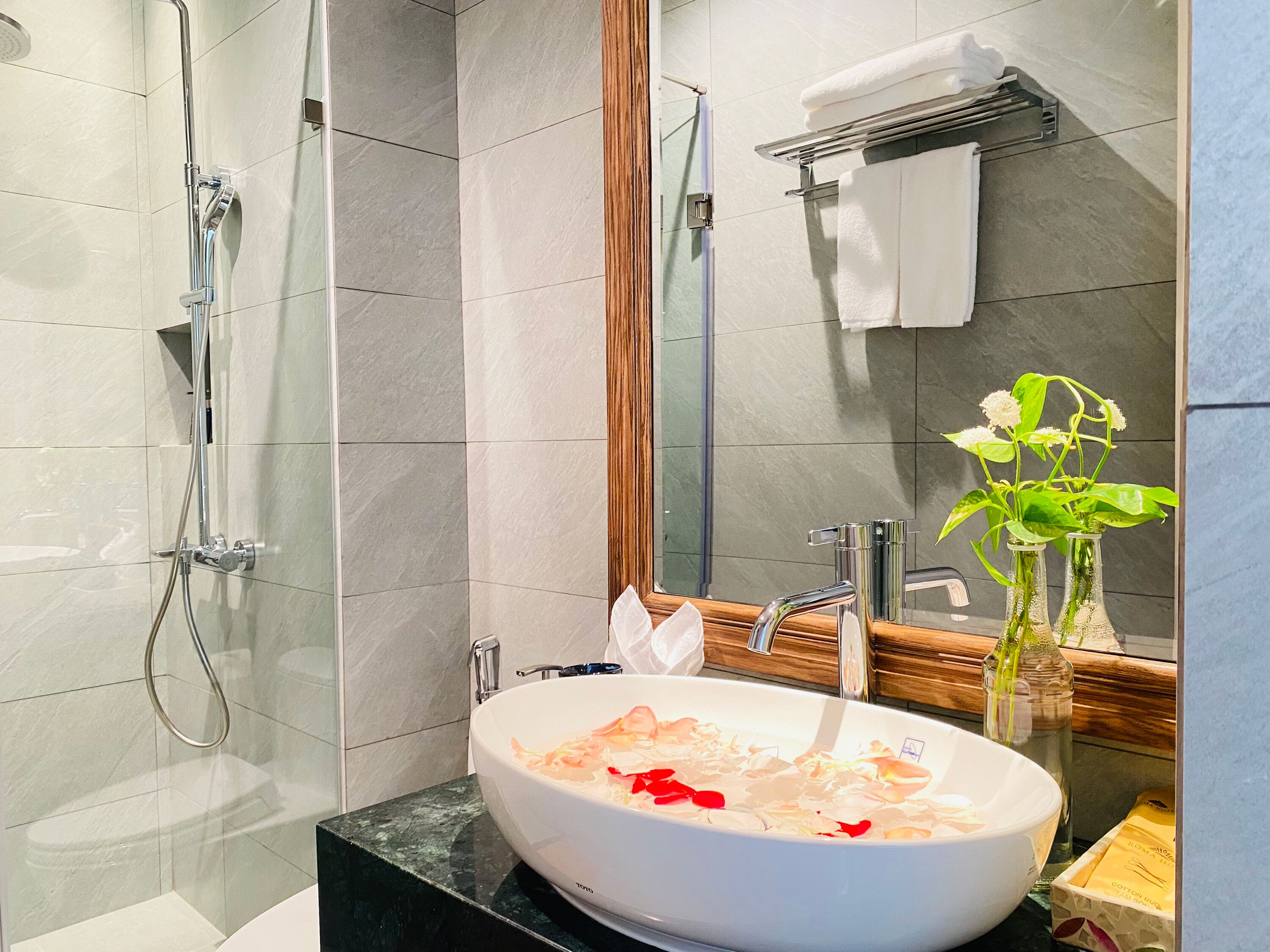 A modern bathroom with a glass-enclosed shower, a white vessel sink with rose petals, a wooden vanity, and a mirror.