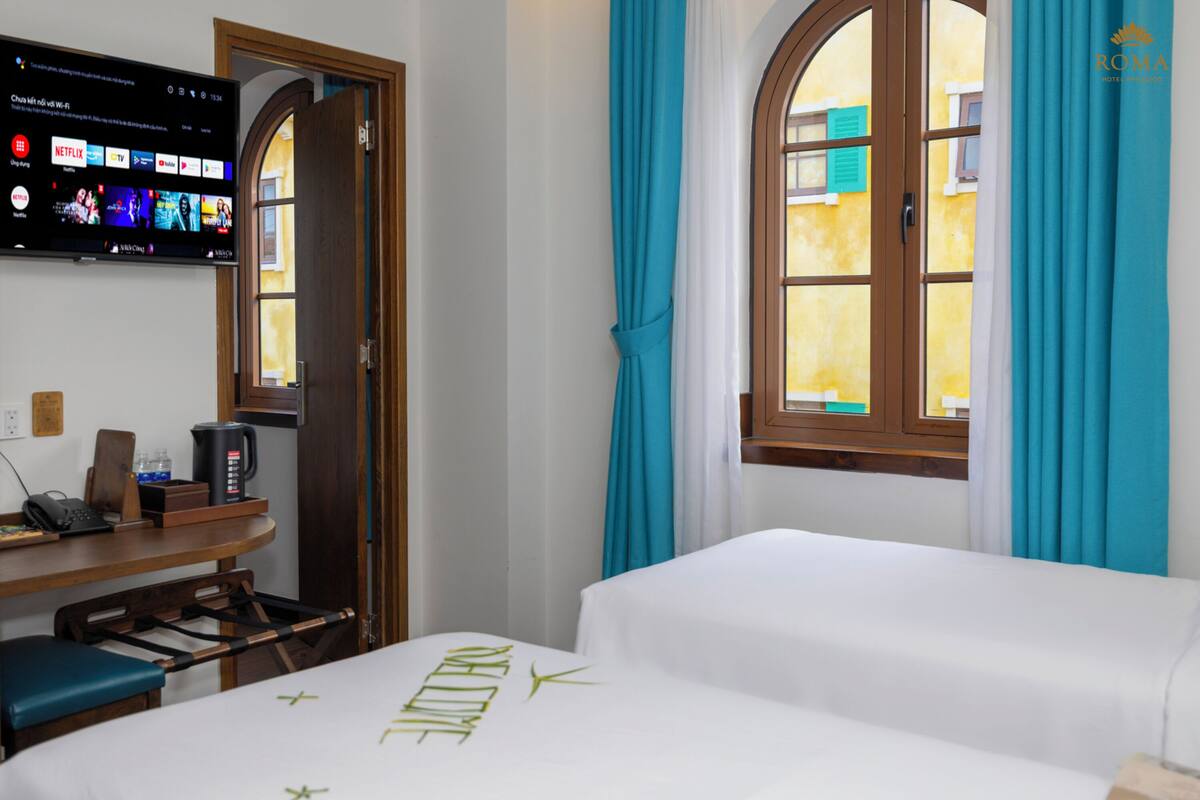 Habitación de hotel con cama, escritorio con cafetera, televisor mostrando servicios de streaming y ventana con cortinas.