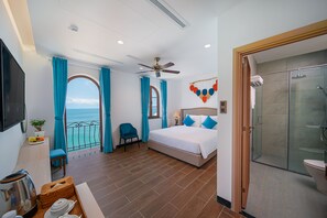 A hotel room with a bed, a chair, a small table with a kettle, a TV, and a view of the sea.