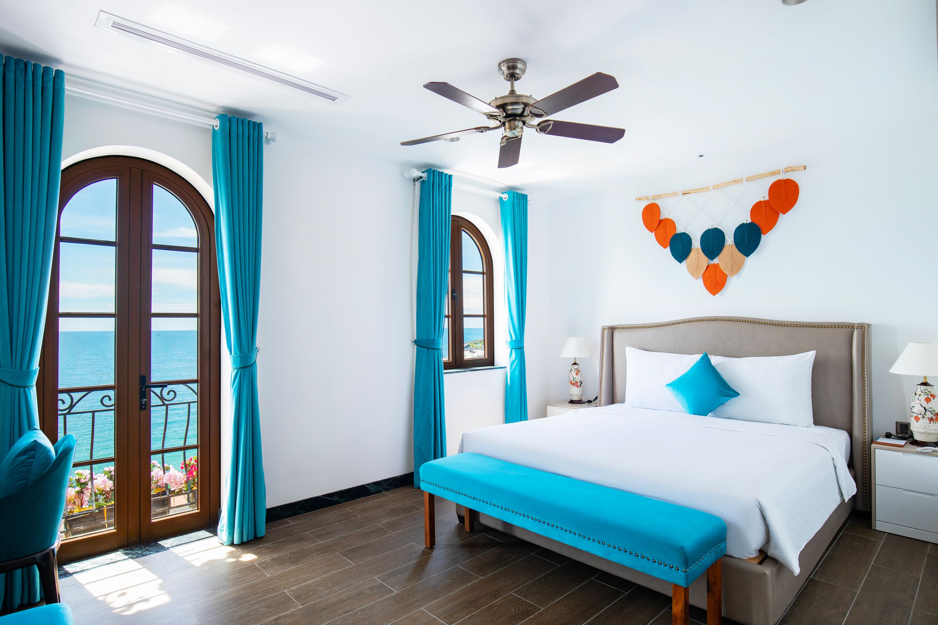 A hotel room with a bed, a nightstand, a ceiling fan, and a view of the sea through a door.