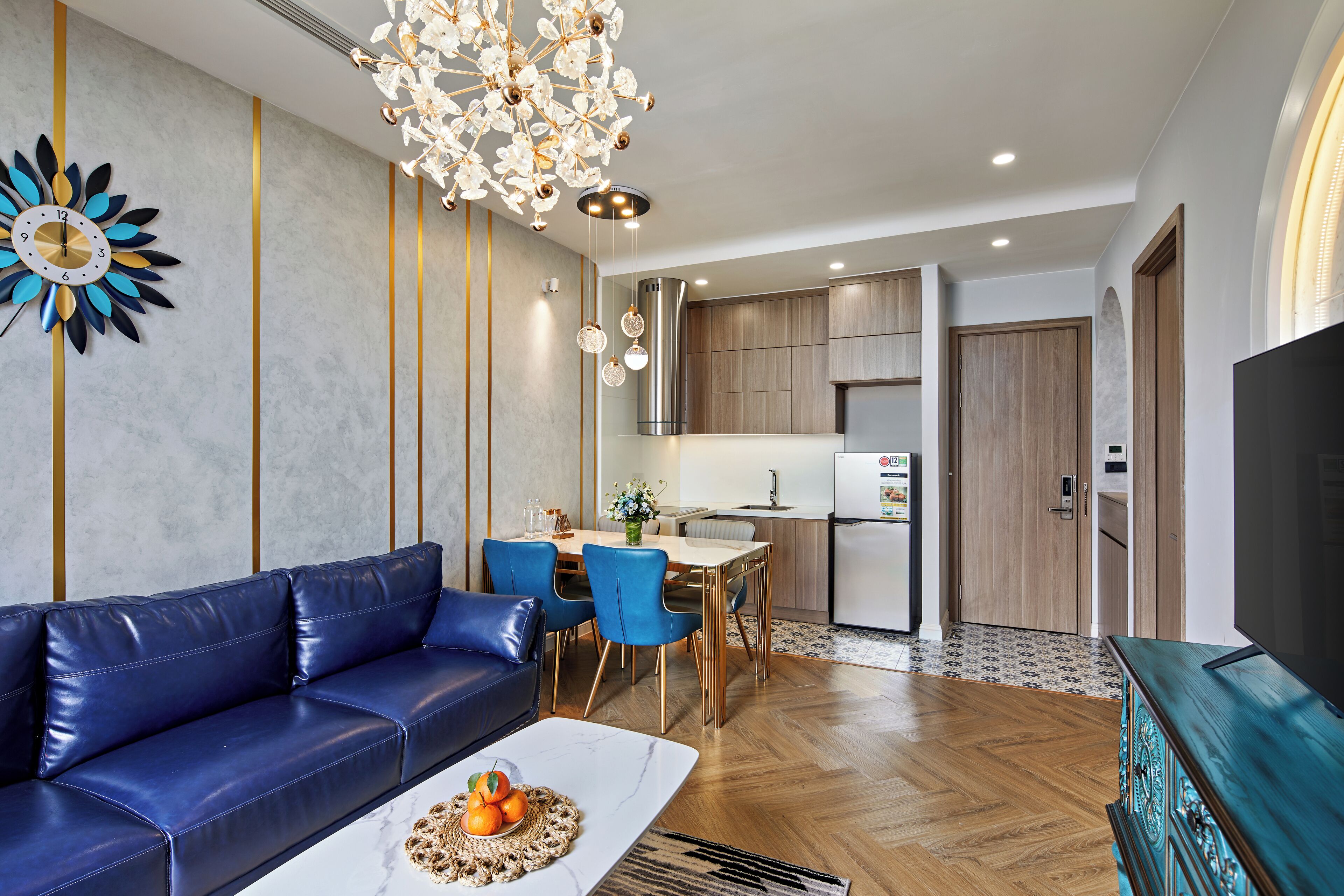 A modern living room with a blue sofa, a dining area with blue chairs, a kitchen with wooden cabinets, and a large flat-screen TV.