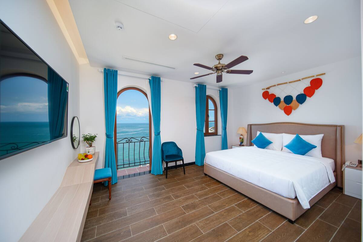 Habitación de hotel con una cama grande, ventilador de techo, vista al mar y balcón con cortinas azules.