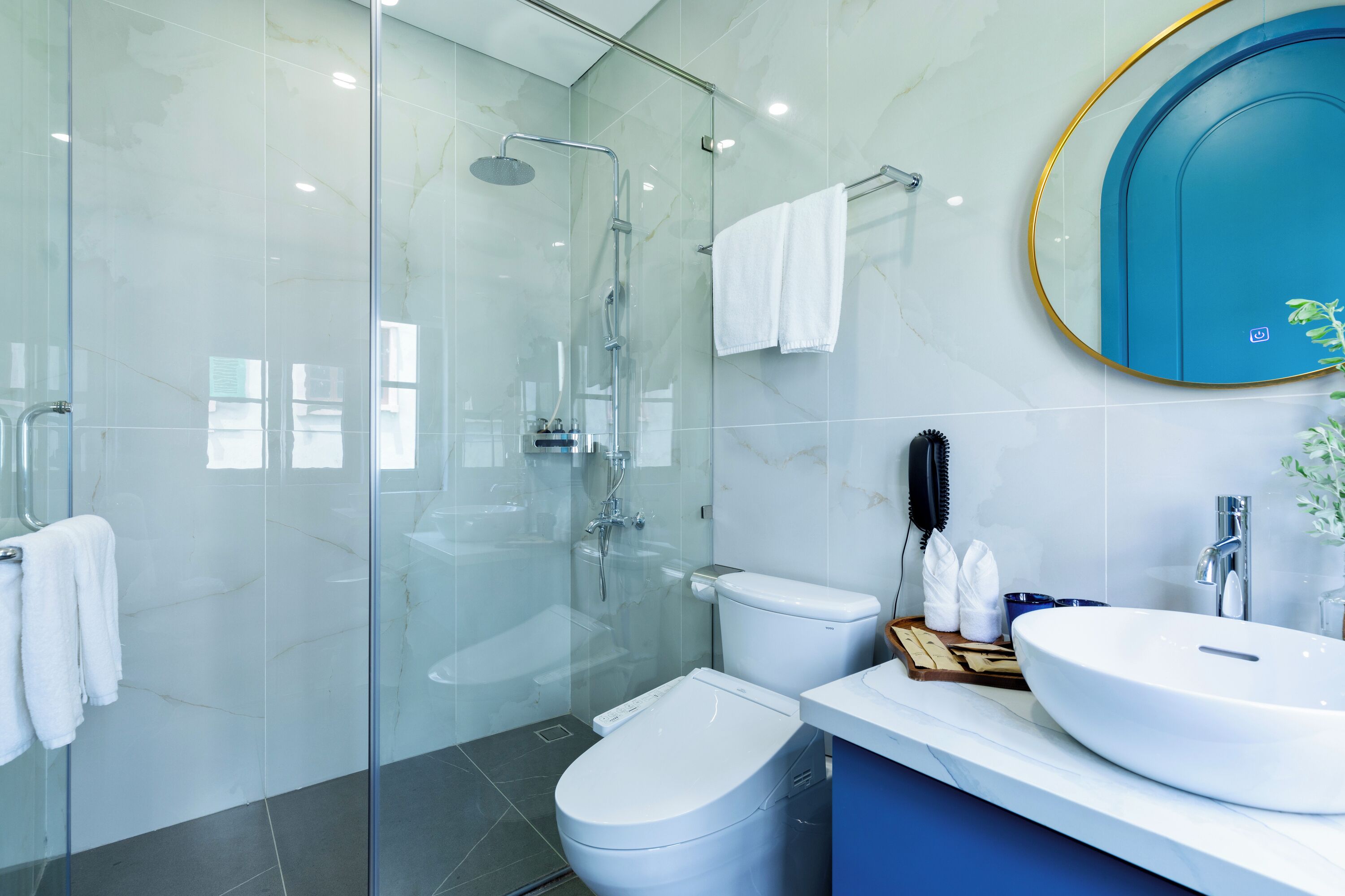 A modern bathroom with a glass-enclosed shower, a white toilet, a sink with a blue countertop, and a blue-framed mirror.