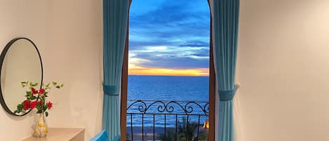 Un balcón con vista al mar al atardecer, un florero con flores y un espejo en la pared.