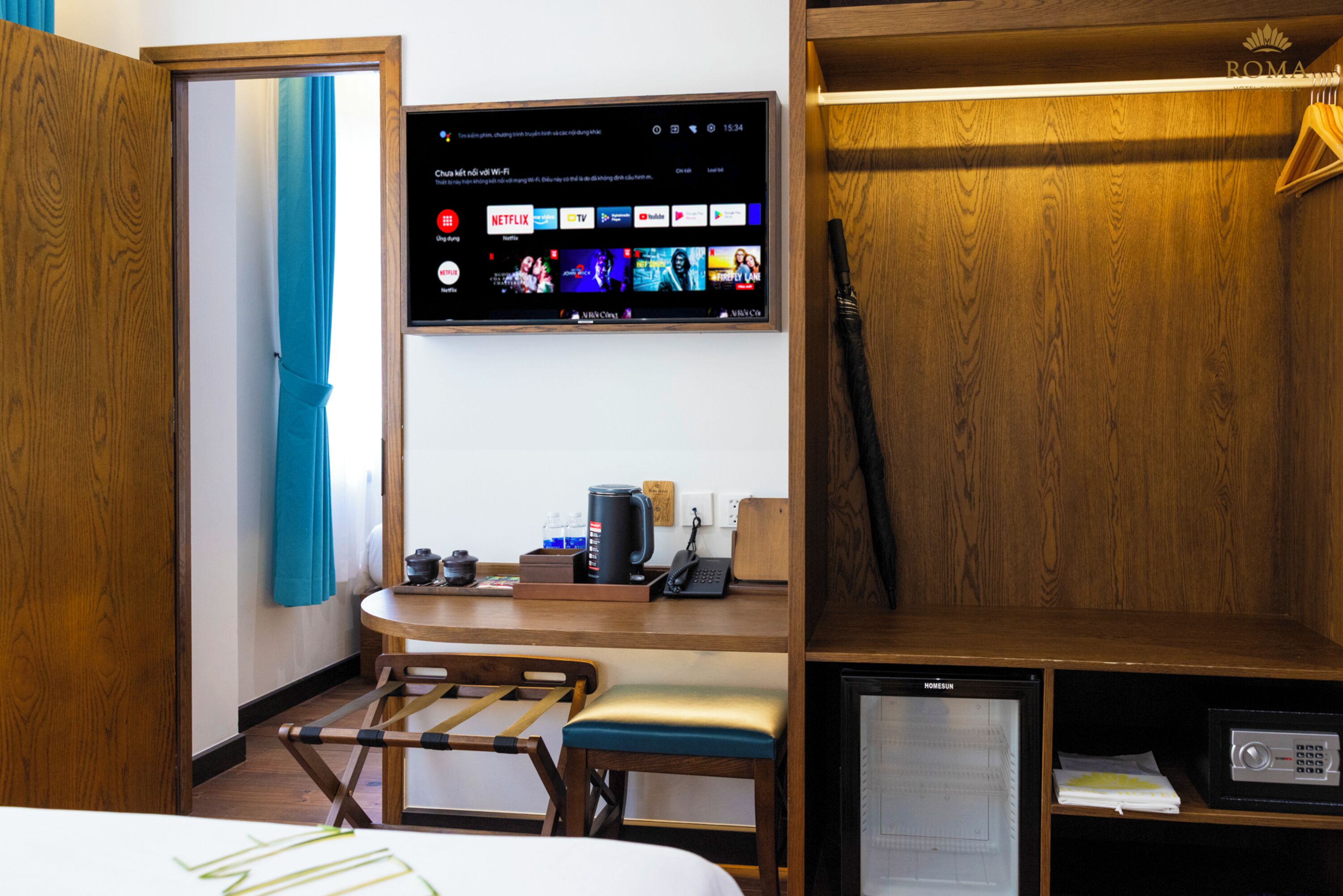 A hotel room with a wooden desk, a small refrigerator, a television mounted on the wall displaying various channels, and a window with blue curtains.