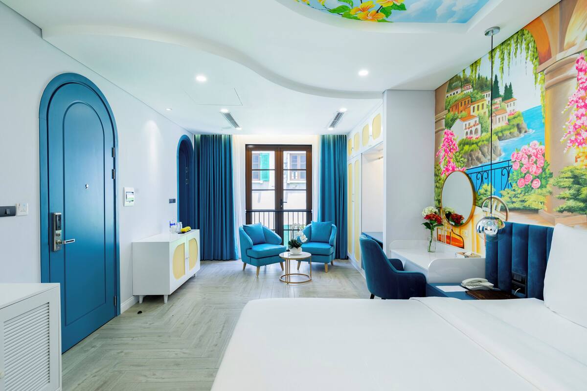 Una habitación de hotel moderna con una cama grande, dos sillones azules, una mesita redonda pequeña, una mesita de noche blanca, un espejo grande y un mural de una escena costera.