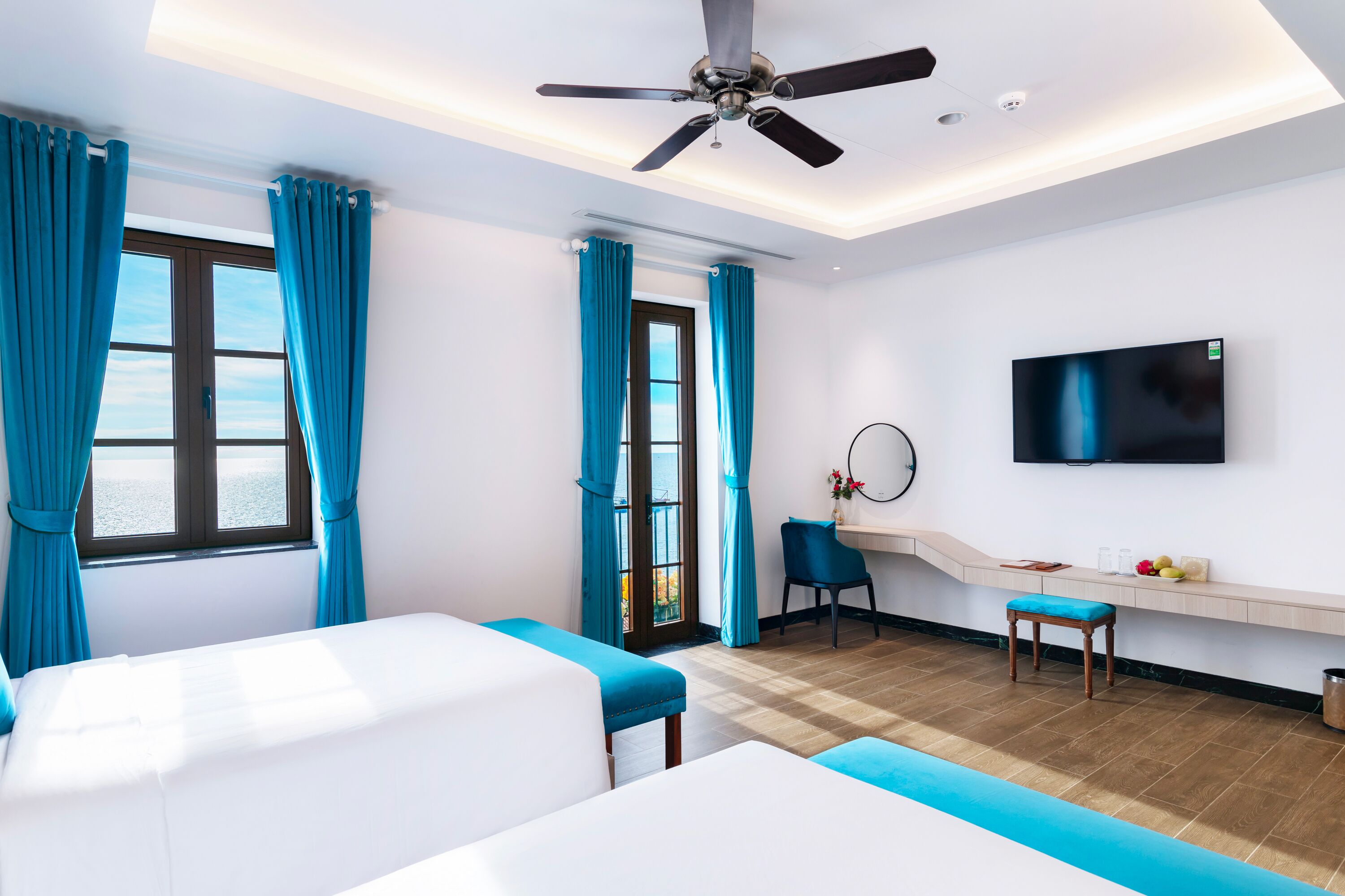 A hotel room with a bed, a desk, a chair, a TV, and a view of the sea.