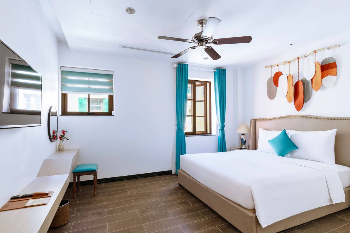 Una habitación de hotel con una cama, un escritorio, una silla, un ventilador de techo y una ventana con cortinas.