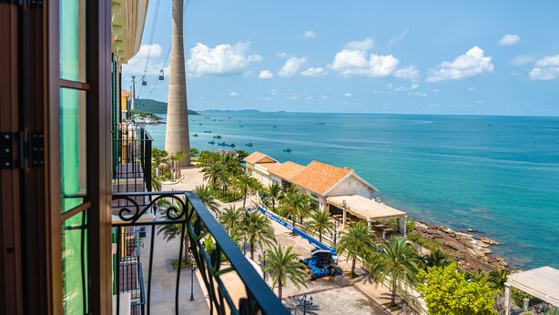 A coastal view from a balcony with a clear sky, a beach, and a building with a red roof.