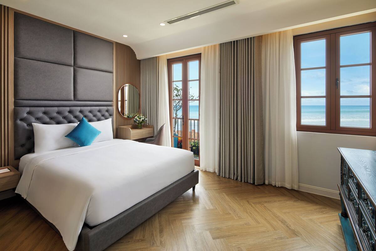 Una habitación de hotel moderna con una cama grande, un cabecero de madera, una mesita de noche, un aparador y vistas al océano.