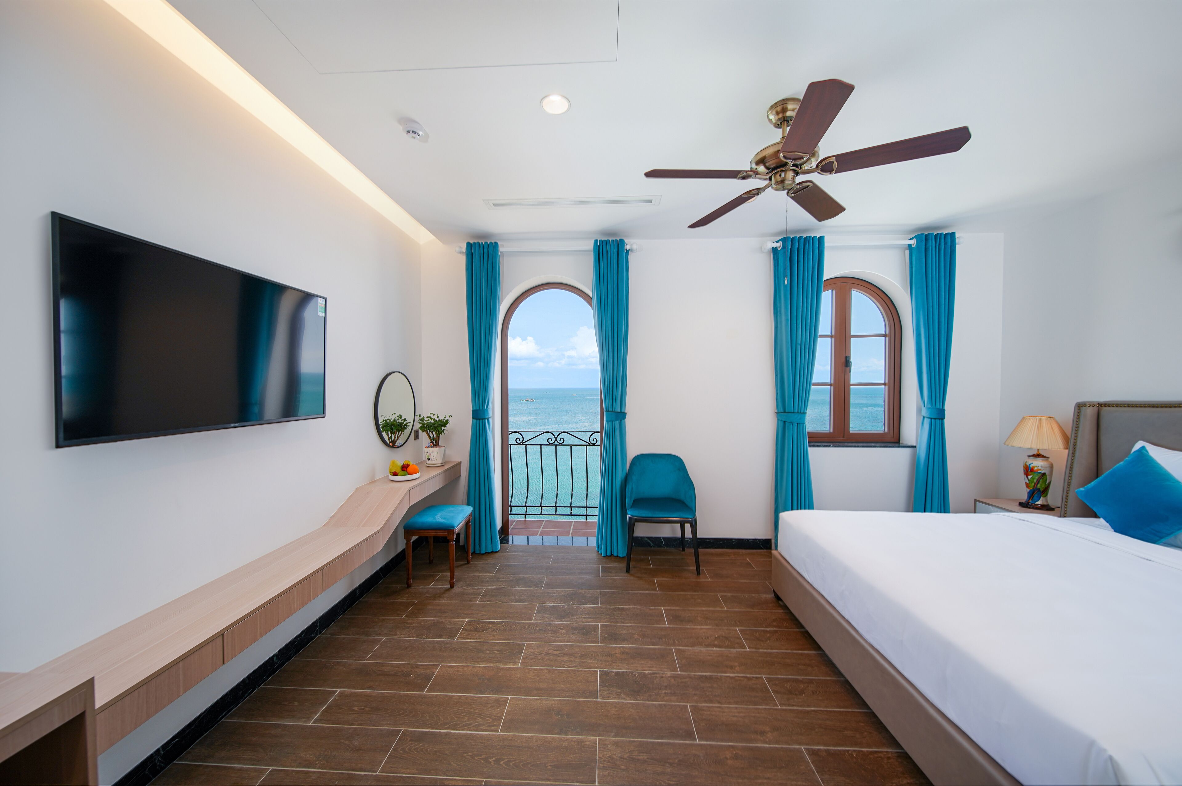 A hotel room with a flat-screen TV, a bed, a desk with a chair, and a balcony with a view of the sea.