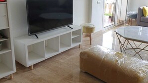 Una sala de estar con una televisión de pantalla plana grande sobre un mueble blanco, una mesa de centro de vidrio y un reposapiés acolchado.