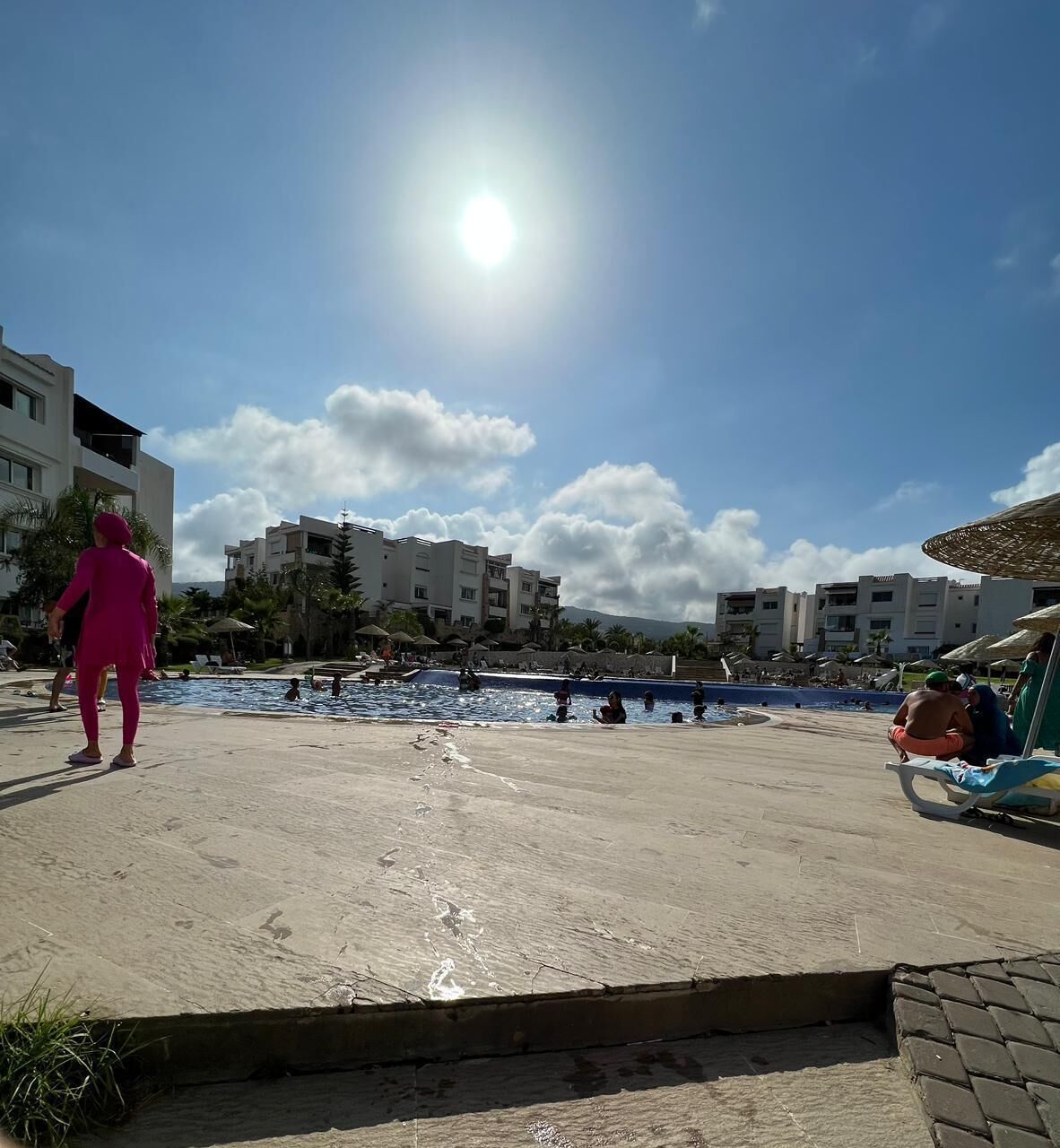 A sunny day at a resort with a swimming pool, palm trees, and buildings in the background.