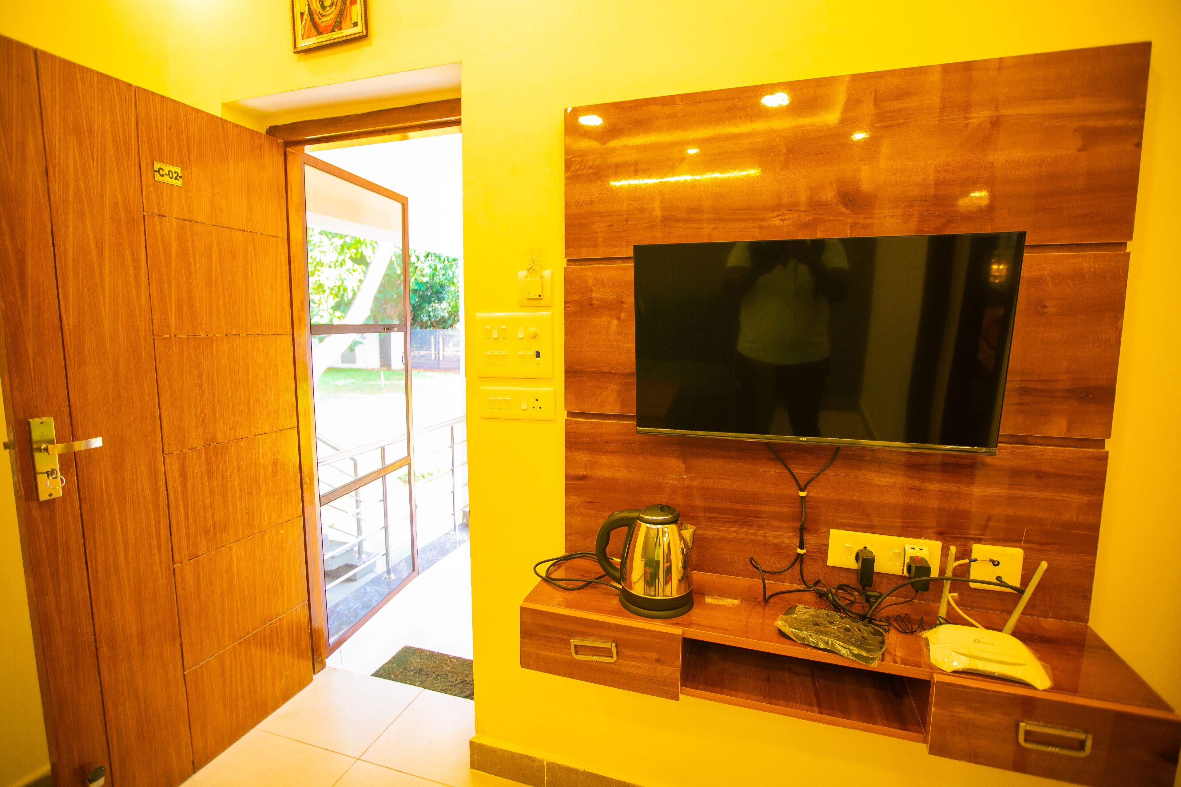 A hotel room with a wooden door, a flat-screen TV mounted on the wall, a coffee maker, and a small table with a power outlet.