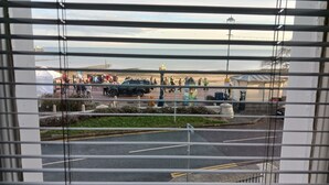 Vista attraverso persiane di una strada sullo sfondo di una spiaggia, una tenda bianca e un veicolo nero.